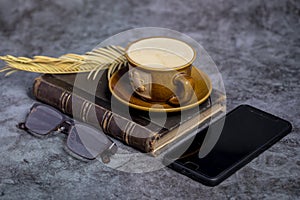 A cup of coffee, an old book , glasses, and a phone on the table