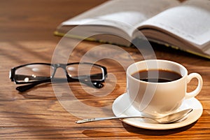 Cup of coffee, open book and glasses on a desk.