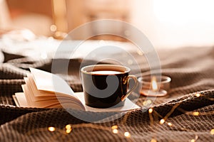 Cup of coffee on open paper book with burning scented candle in bed with glow lights at home.