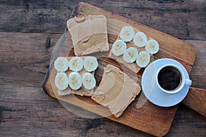 Cup of coffee and peanut butter banana toasts on wooden background. Slices of whole wheat bran bread with peanut paste on cutboard