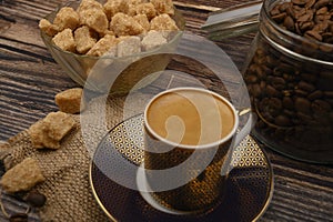 A Cup of coffee, pieces of brown sugar in a sugar bowl, coffee beans in a glass jar on a wooden background