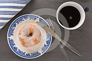 A cup of coffee, a plate with sugar coated doughnut and other breakfast objects on tabletop
