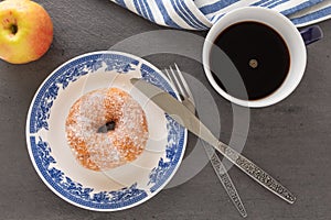 A cup of coffee, a plate with sugar coated doughnut and other breakfast objects on tabletop