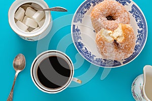 A cup of coffee, a plate with sugar coated doughnuts and other breakfast objects, on blue background