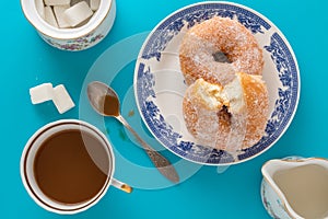 A cup of coffee, a plate with sugar coated doughnuts and other breakfast objects, on blue background