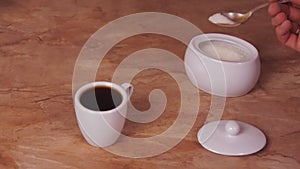The cup of coffee poured sugar. The white cup of hot coffee with sugar added. Marble Table. White sugar bowl.