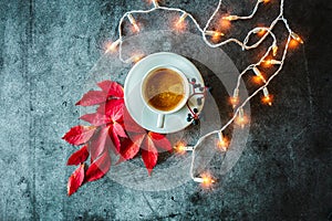 Cup of coffee, red autumn leaves, berries, glowing garland on a concrete background