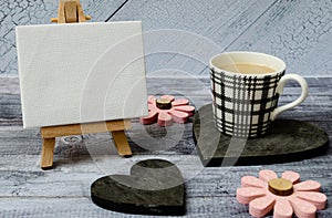 Cup of coffee, small easel with a blank canvas over white, two dark wooden hearts and pink flowers. Old wooden