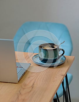 a cup of coffee stands on a table in a cafe near a laptop against the background of a turquoise chair. distant work.