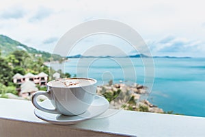 A cup of coffee stands on the window with a beautiful view of the ocean on the tropical island