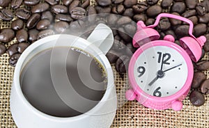 Cup of coffee with steam and pink clock on table with coffee beans background. Concept coffee and time