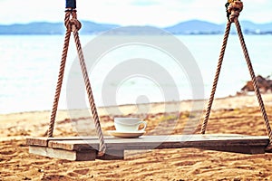 A Cup of coffee is on the swing in the morning near the beach.