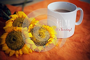 Cup of coffee or tea and flowers arrangement on orange background. With self reminder text on white mug - Relax. It is Sunday.