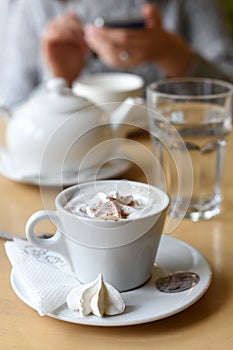 A cup of coffee, a tea pot and a pair of woman`s hands holding a mobile phone in the background