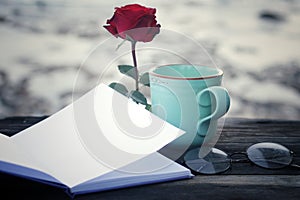 A cup of coffee or tea and red rose with blank paper book on bright background. Notebook, red rose flower, glasses and coffee cup.