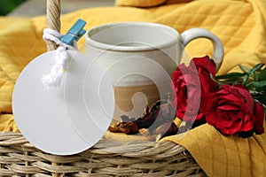 Cup of coffee or tea and red rose flowers in a rattan basket on yellow background. With white blank tag label paper for your text.