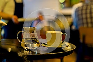 Cup of coffee or tee, glass teapot of hot water, morning. Image with blurred cafe visitors and a waiter in a low key