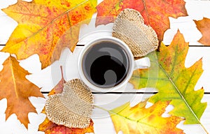 Cup of coffee with two burlap hearts and autumn leaves on white wooden table.Top view