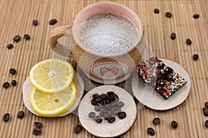 A cup of coffee on a wooden stand. Nearby lies a yellow juicy lemon, dark chocolate with fruits and coffee beans scattered in the