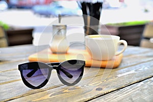 Cup of coffee on a wooden stand is on the table in a cafe summer morning, sunglasses in the foreground