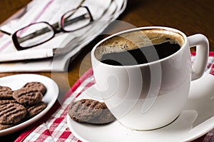 Cup of coffee on a wooden table with biscuits, glasses and newspaper