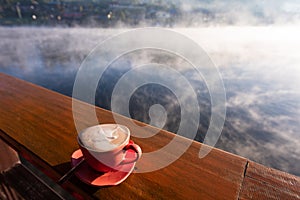 A cup of coffee on wooden table with steam over lake at Rak thai village