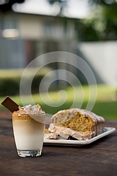 Cup of Dalgona coffee served on a glass with a bar of chocolate and a wholemeal cake. Outdoor background