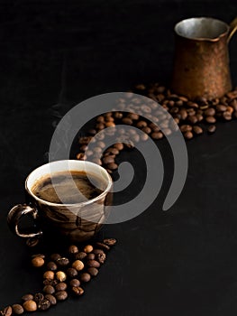 A cup with dark espresso on a black background, steam rises above the cup. Roasted coffee beans are located around a cup of coffee