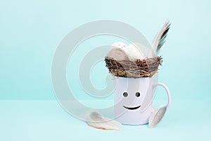 Cup with an easter egg nest, bird feathers and a smiling happy face, spring holiday greeting card