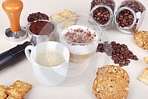 Cup of espresso, cappuccino with chocolate crumbs, cookies, crackers, holder with ground coffee, tamper and coffee beans on table