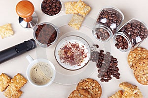 Cup of espresso, cappuccino, crackers, cookies, holder with ground coffee, tamper and coffee beans on table. View from above
