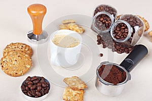 Cup of espresso, crackers, cookies, holder with ground coffee, tamper and cans of coffee beans on table