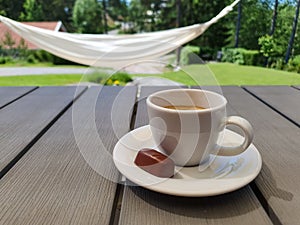 Cup of espresso with small chocolate praline on gray table with hammock and green sunny garden in the background