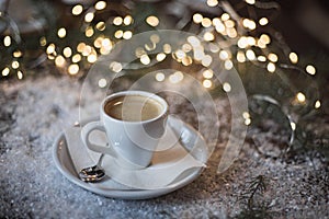 A cup of espresso surrounded by New Year`s lights and pine branches in the snow. Dark background. Christmas mood