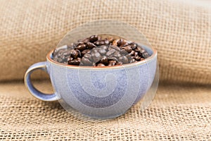 Cup filled with brown roasted coffee beans over folded burlap background