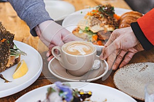 a cup of flat white coffee in cream ceramic cup with latte art on top, touched by two hands, surrounded with variety of food on