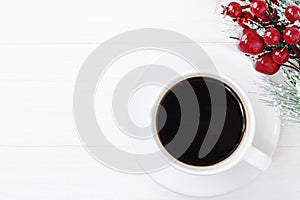 Cup of fresh coffee on a white wooden background next to a red snowy Christmas branch. Christmas, New Year`s concept. Top view,