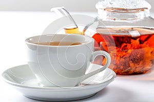 A Cup of fresh tea on a saucer, behind the Tea in a glass teapot with a blooming large flower. Exotic green tea on white