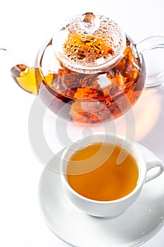 A Cup of fresh tea on a saucer, behind the Tea in a glass teapot with a blooming large flower. Exotic green tea on white