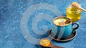 Cup of golden turmeric milk, wooden spoon with curcuma powder and jar of honey on blue. Selective focus. Copy space