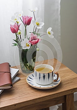 A cup of green tea, a bouquet of spring garden flowers, a notebook on a wooden table in a cozy living room. Slow tea party