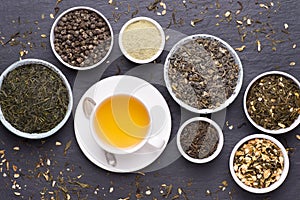Cup of green tea and bowls of various dried tea leaves on dark, stone background