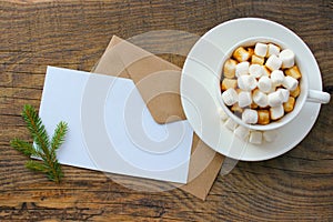 Cup with homemade Christmas hot chocolate drink, marshmallows and holiday greeting card on wooden background.