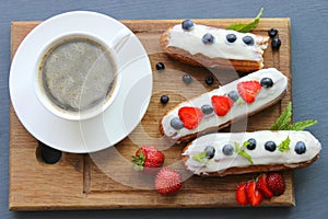 A Cup of hot black coffee and delicious eclairs with strawberries and blueberries on a wooden cutting Boar