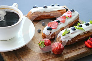 A Cup of hot black coffee and delicious eclairs with strawberries and blueberries on a wooden cutting Boar