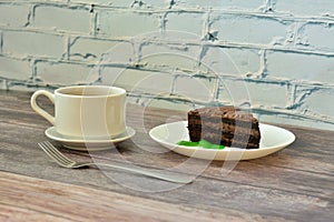 A cup of hot black coffee on a saucer with a spoon and a plate of layered chocolate cake on a wooden table against a brick wall