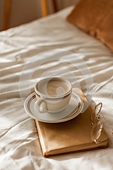 A cup of hot cappuccino, a book, glasses on the bed. Breakfast. Cozy house. Decor, details