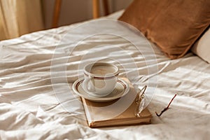 A cup of hot cappuccino, a book, glasses on the bed. Breakfast. Cozy house. Decor, details