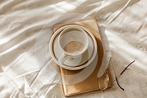 A cup of hot cappuccino, a book, glasses on the bed. Breakfast. Cozy house. Decor, details