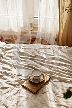 A cup of hot cappuccino, a book, glasses on the bed. Breakfast. Cozy house. Decor, details